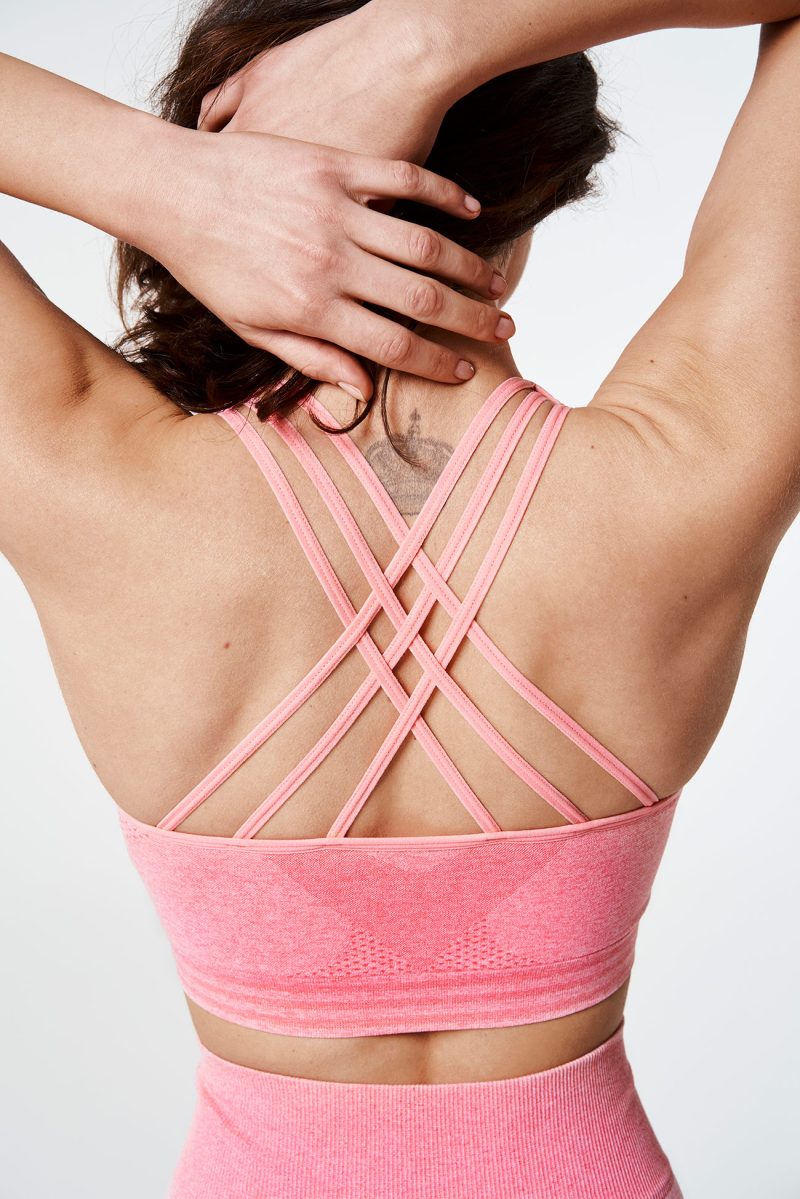 Close-up of a brunette woman’s back, wearing a Coral Pink Bold Seamless Performance Strappy Back Sports Bra, with her hands holding her hair up, emphasizing the strappy design and smooth fit.