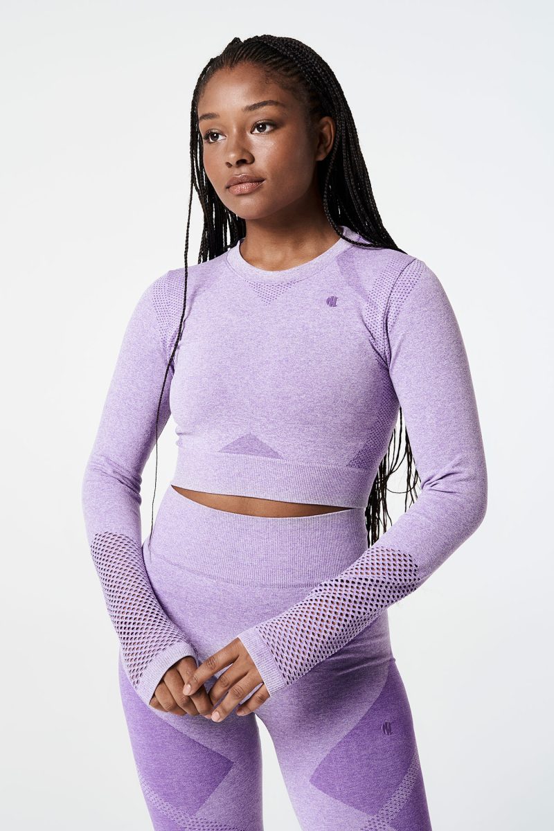 A Black woman facing forward, wearing a violet pink bold seamless performance long-sleeve crop top with mesh details on the arms and matching shorts, displaying a confident and stylish athletic look.