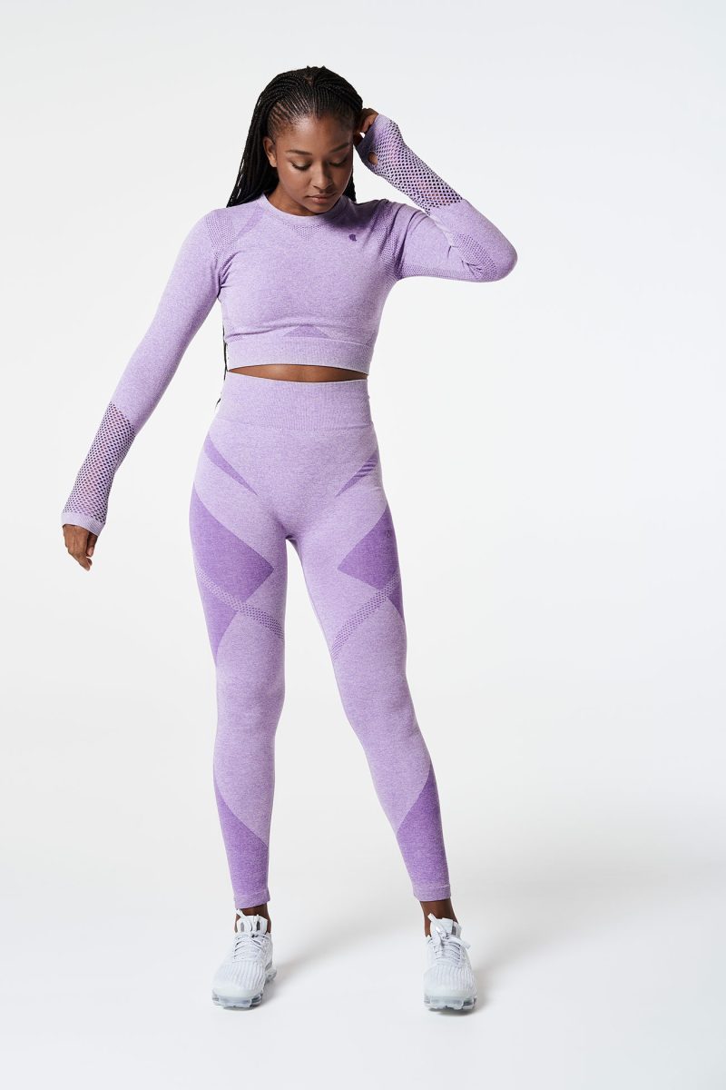 Full-body shot of a Black woman facing forward and looking down, wearing a violet pink bold seamless performance long-sleeve crop top with mesh details on the arms and matching leggings, highlighting the sleek and athletic design.